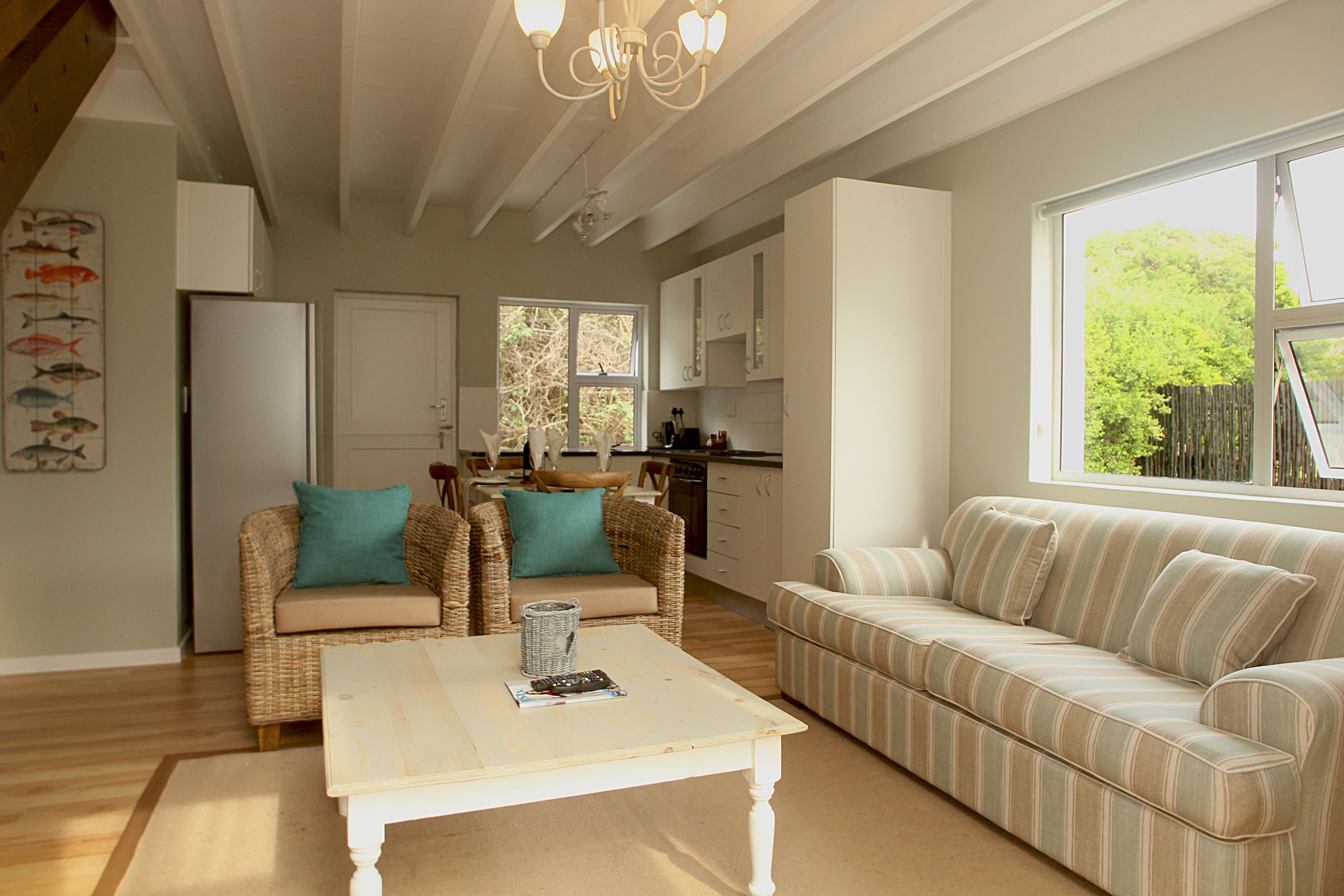 A spacious living area with a white coffee table, two sofas, a kitchen in the background, and large windows offering a view of greenery.
