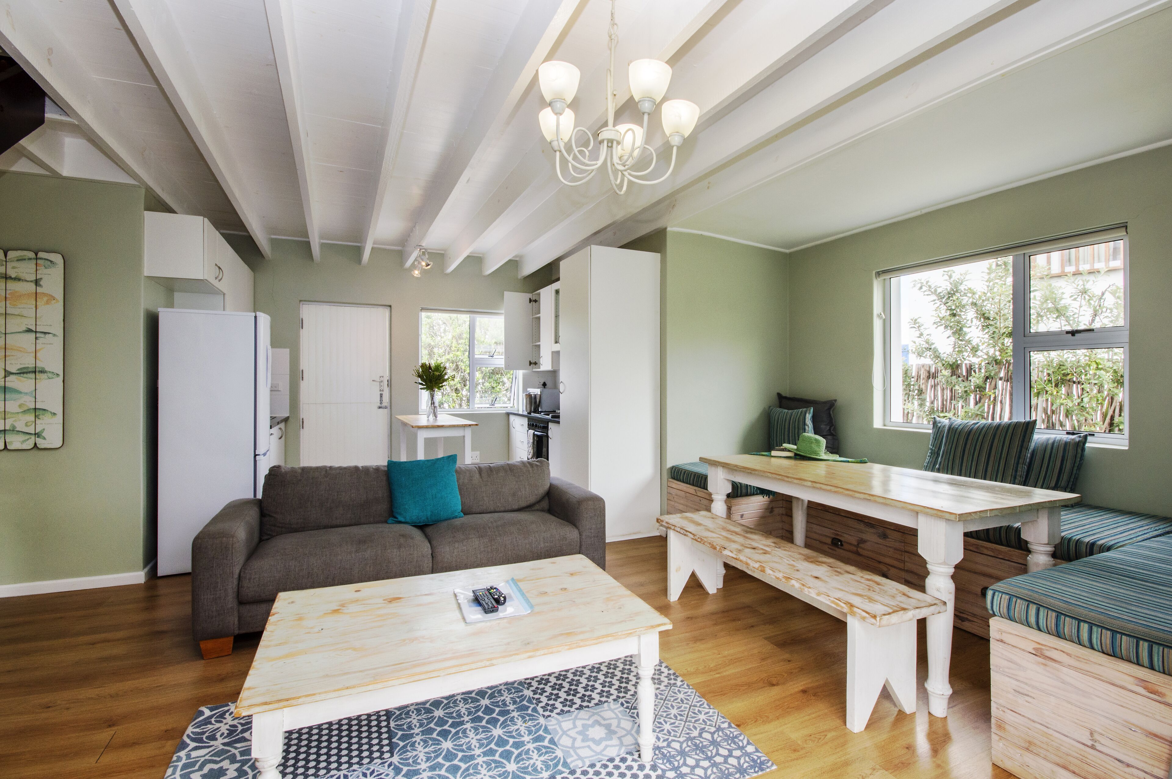 Amplia sala de estar con una mesa de madera, un sofá y un banco. Se observa una kitchenette al fondo y una ventana con vista a la vegetación.