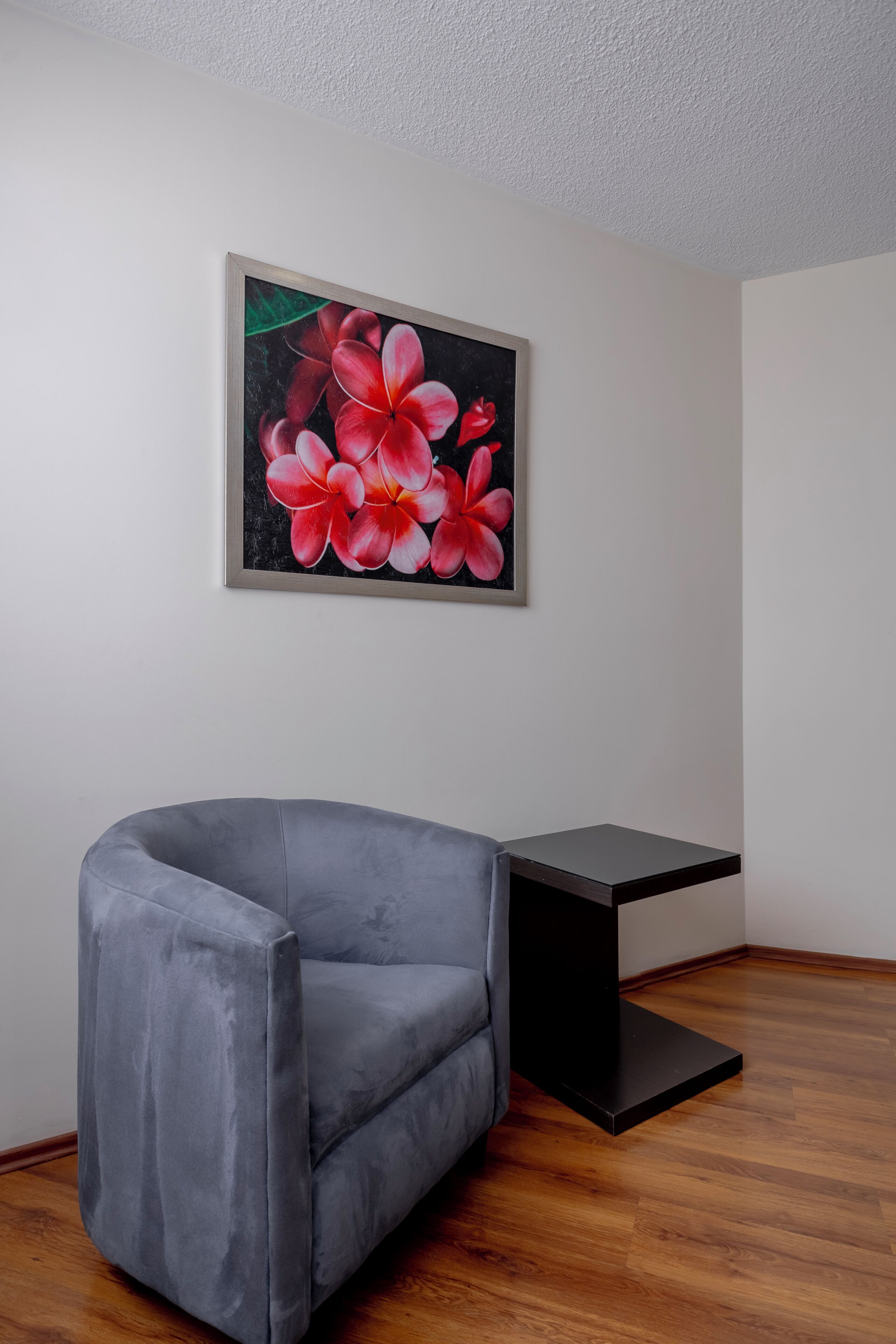 Ein Zimmer mit einem grauen Sessel, einem kleinen schwarzen Tisch und einem gerahmten Bild von roten Blumen an der Wand.