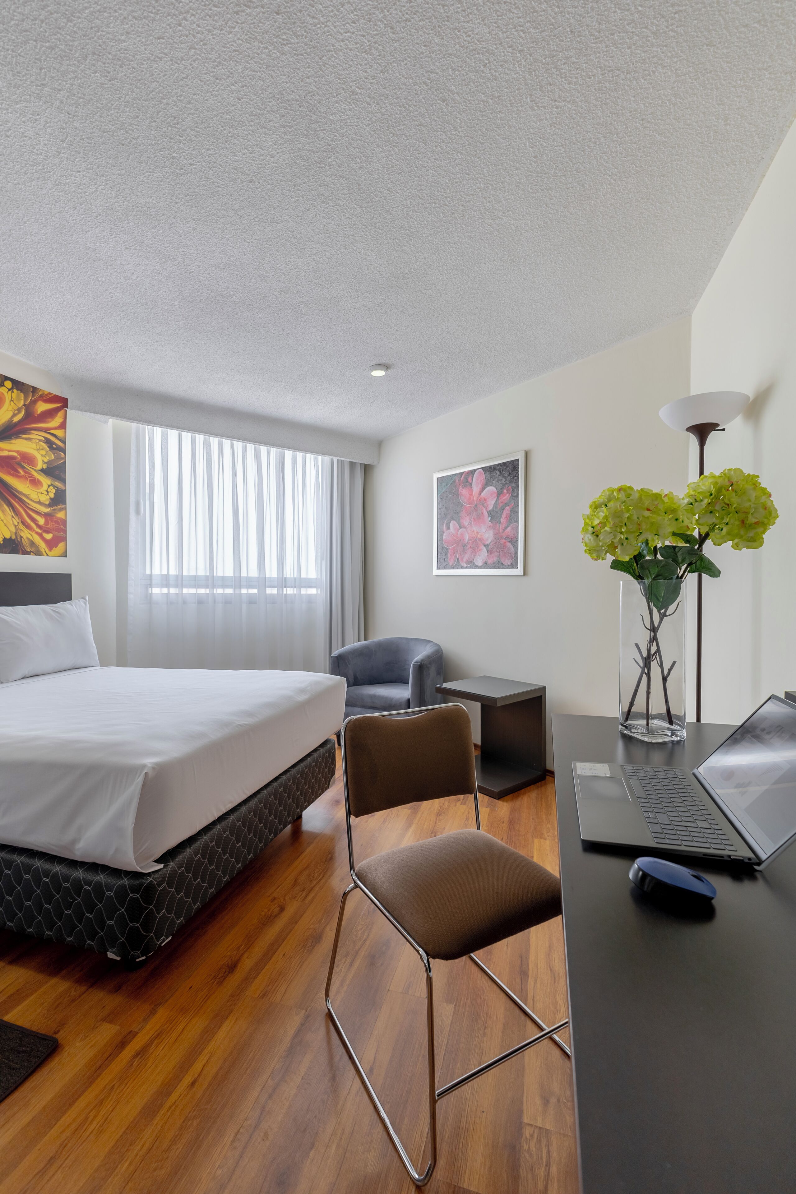 Habitación de hotel con cama, escritorio con computadora portátil, una silla, una mesita, un florero con flores y obras de arte en las paredes.