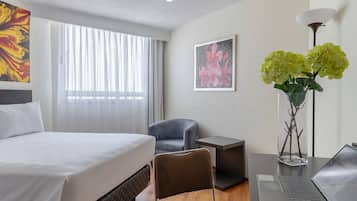 Habitación de hotel con cama, escritorio con computadora portátil, una silla, una mesita, un florero con flores y obras de arte en las paredes.