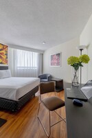 Habitación de hotel con cama, escritorio con computadora portátil, una silla, una mesita, un florero con flores y obras de arte en las paredes.