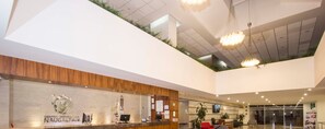 Una recepción de hotel moderna con techo de vidrio, un mostrador de recepción y una pantalla digital.