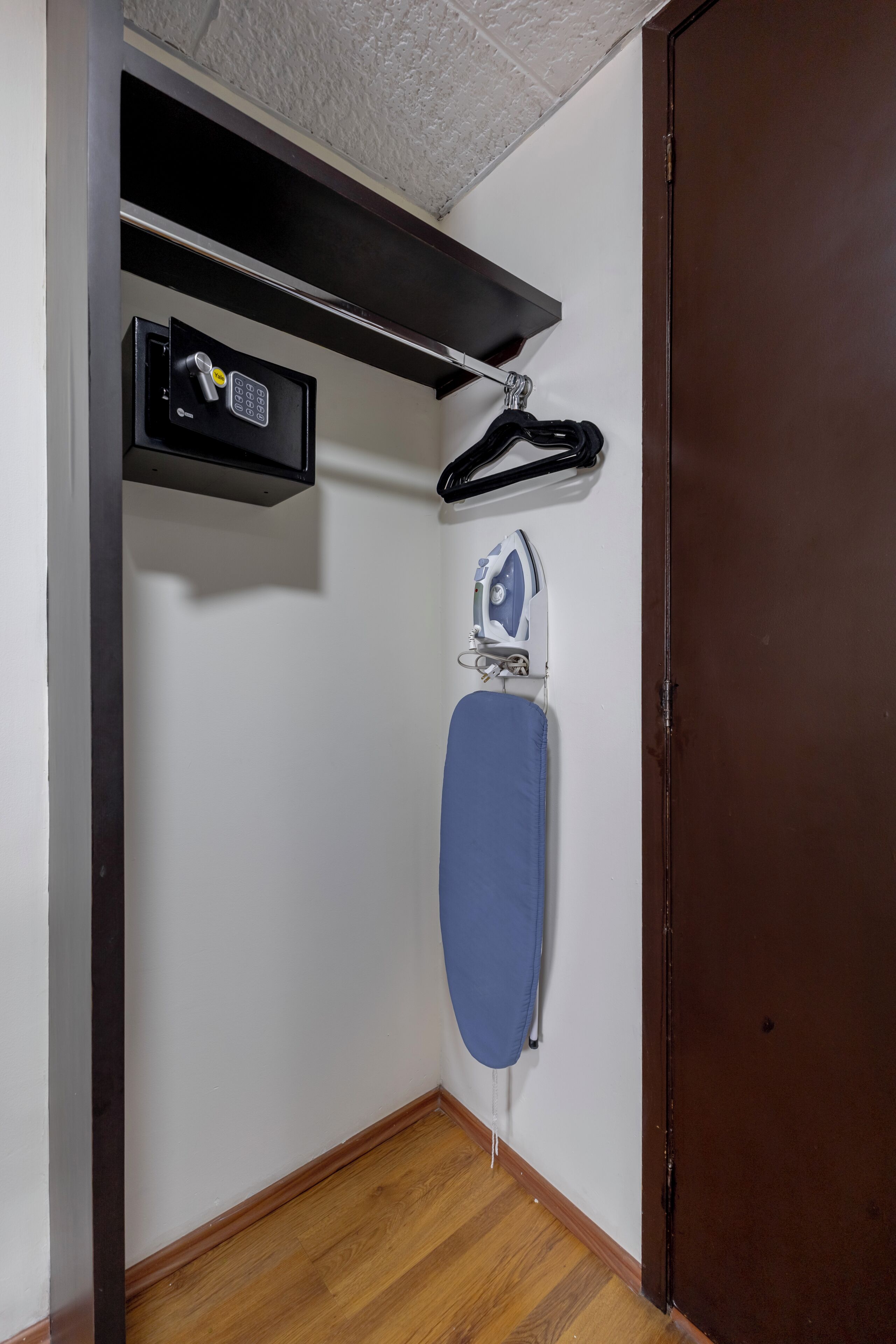 Una habitación pequeña con suelo de madera, pared blanca, un perchero negro con algunos objetos, tabla de planchar azul y puerta marrón.