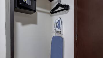 Una habitación pequeña con suelo de madera, pared blanca, un perchero negro con algunos objetos, tabla de planchar azul y puerta marrón.
