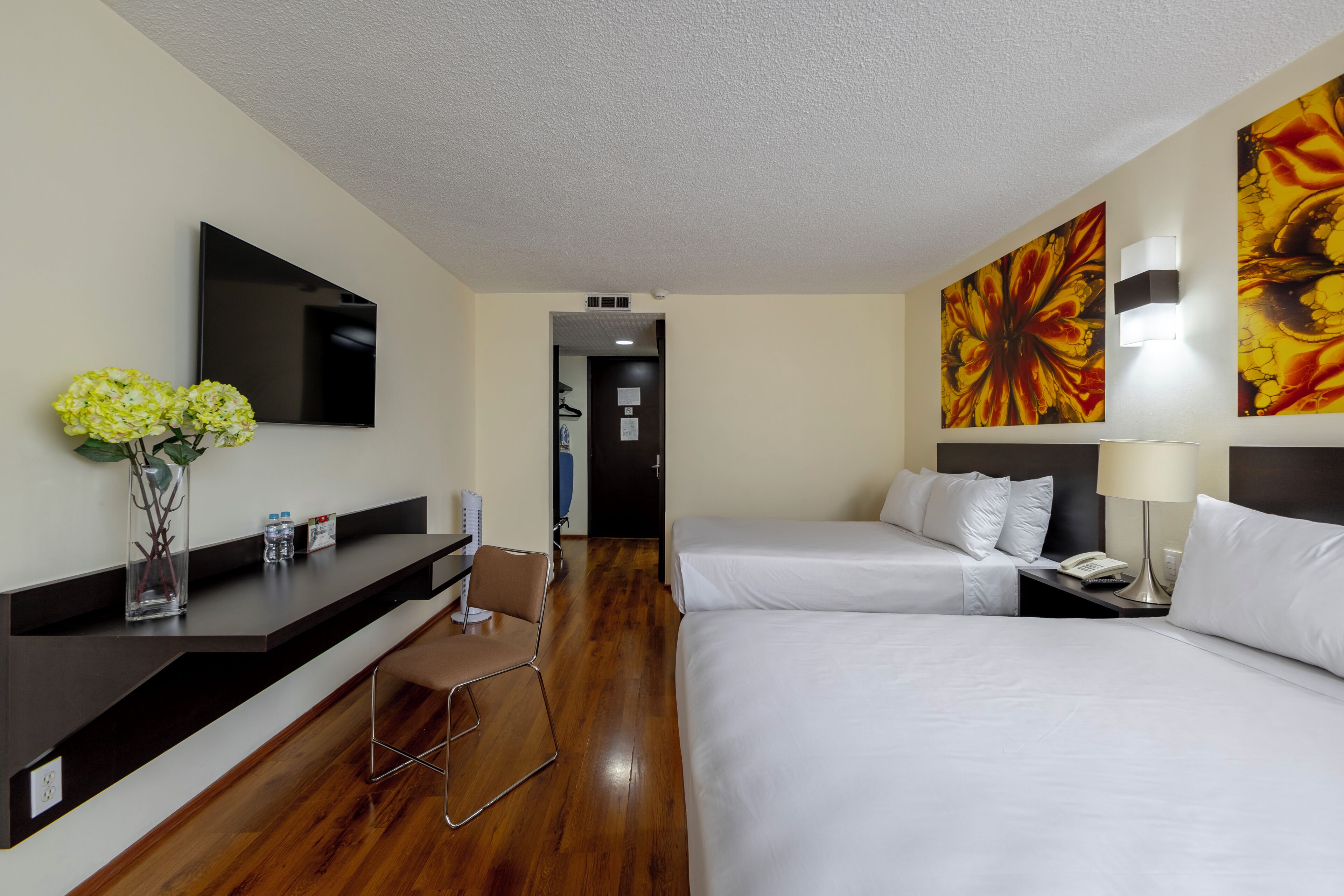 Habitación de hotel con televisión de pantalla plana, un florero con flores, un escritorio con silla y dos camas con ropa blanca.