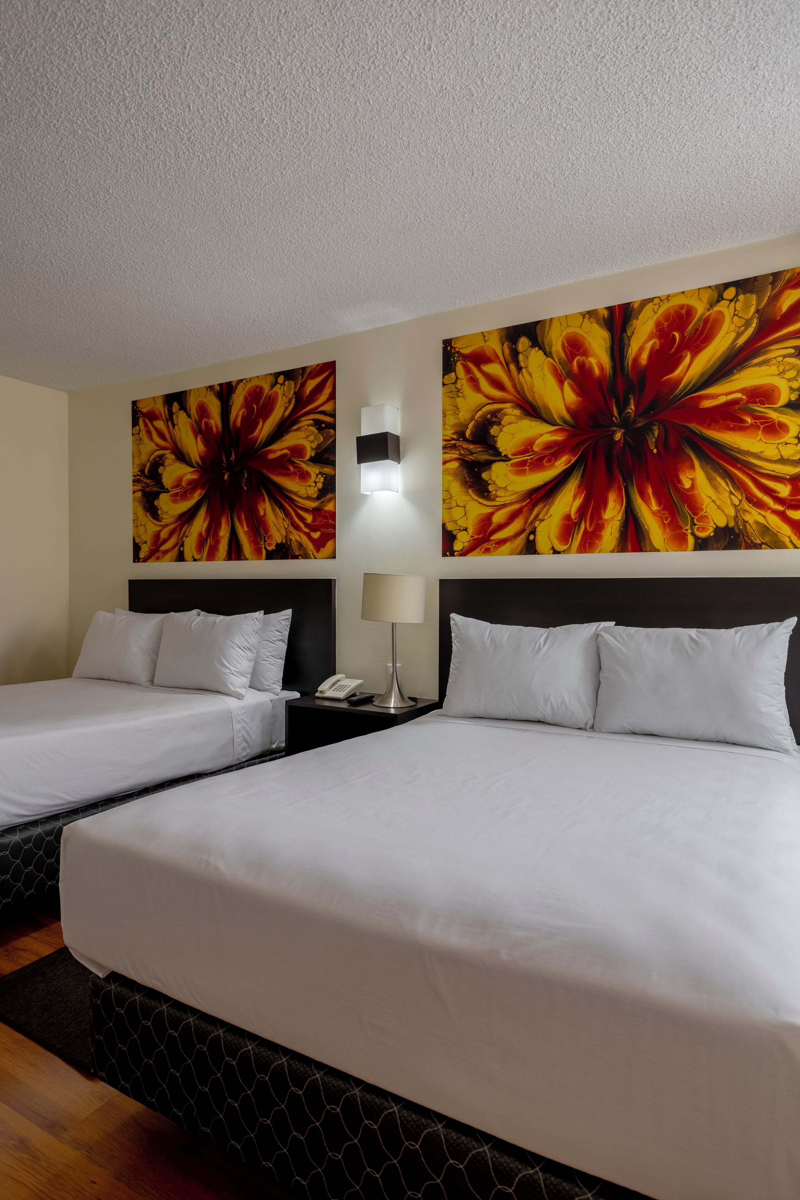 A hotel room with two beds, a nightstand, a lamp, and two large floral paintings on the walls.