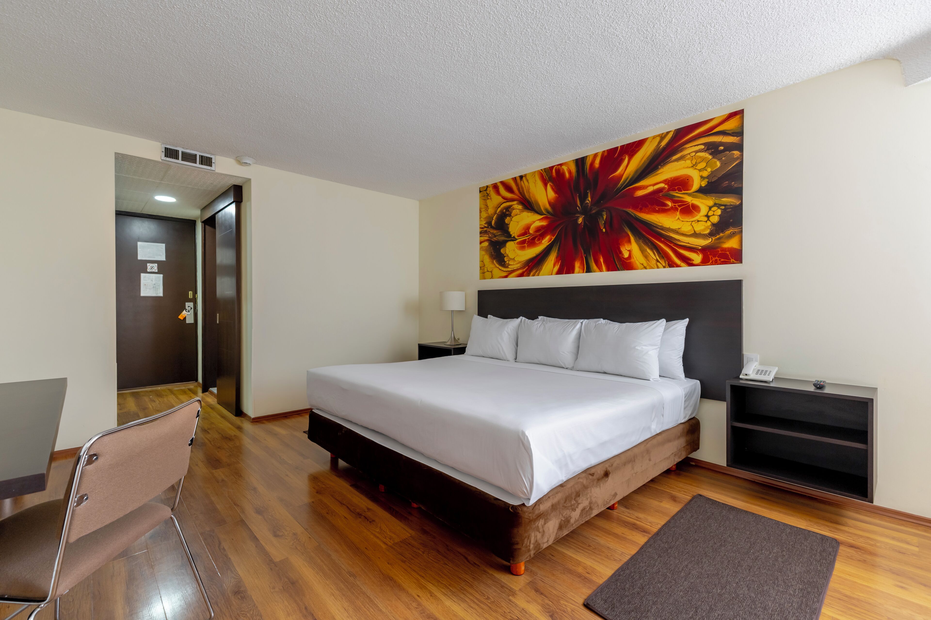 Habitación de hotel con una cama grande, una mesita de noche, un escritorio, una silla, un cuadro en la pared y suelo de madera.