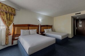 Habitación de hotel con dos camas individuales, cabecera de madera, mesita de noche con lámpara y ventana con cortinas.