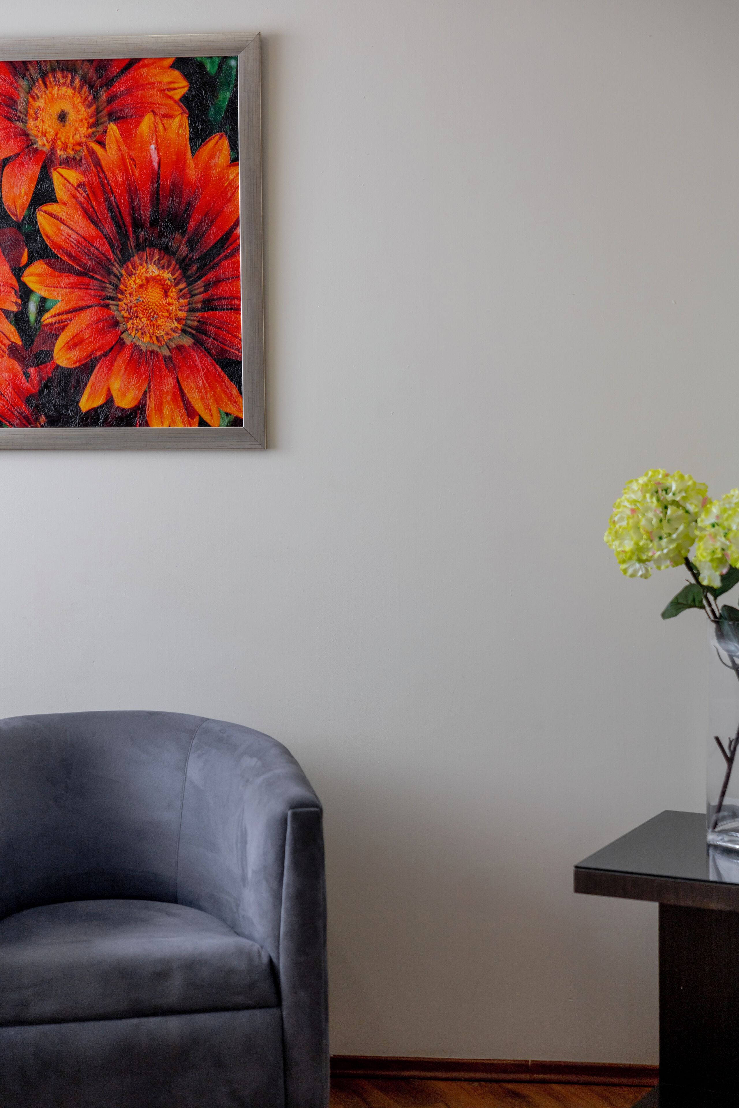 Una habitación con un sillón gris, un cuadro de flores y un florero con flores amarillas sobre una mesa.