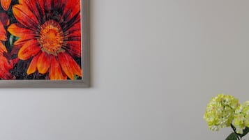 Una habitación con un sillón gris, un cuadro de flores y un florero con flores amarillas sobre una mesa.