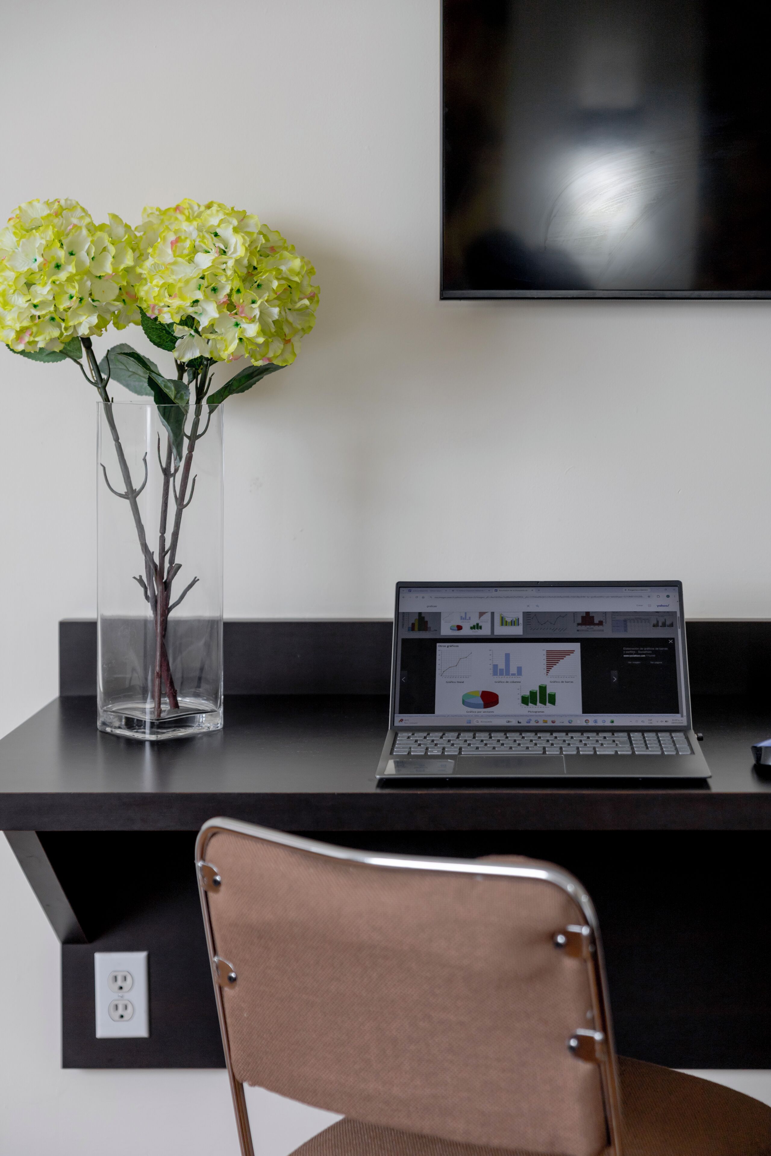 Un espace de travail avec un ordinateur portable affichant des graphiques, un vase avec des fleurs et une chaise.