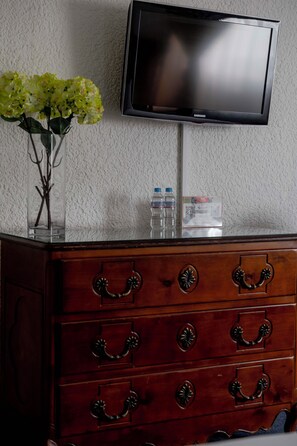 Un aparador de madera con tiradores ornamentales, un jarrón con flores, dos botellas de agua y un televisor montado en una pared con textura.