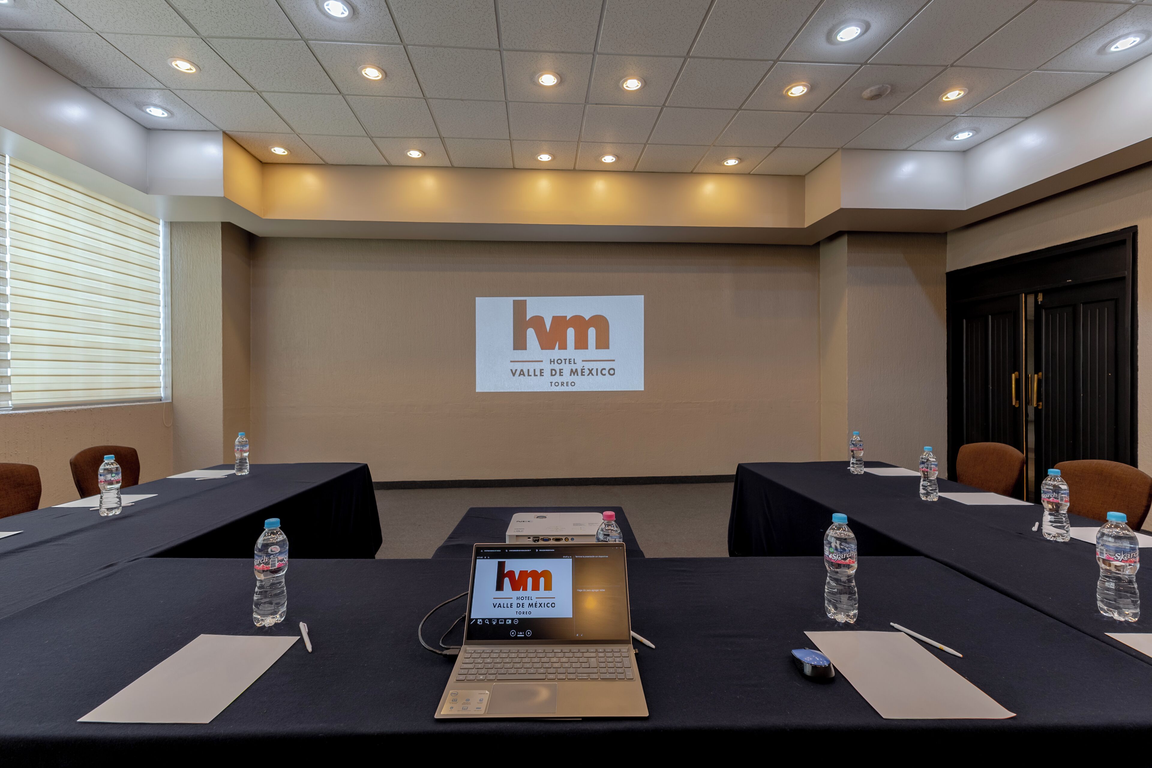 A conference room with a large screen displaying the hotel name, tables with water bottles, and laptops.
