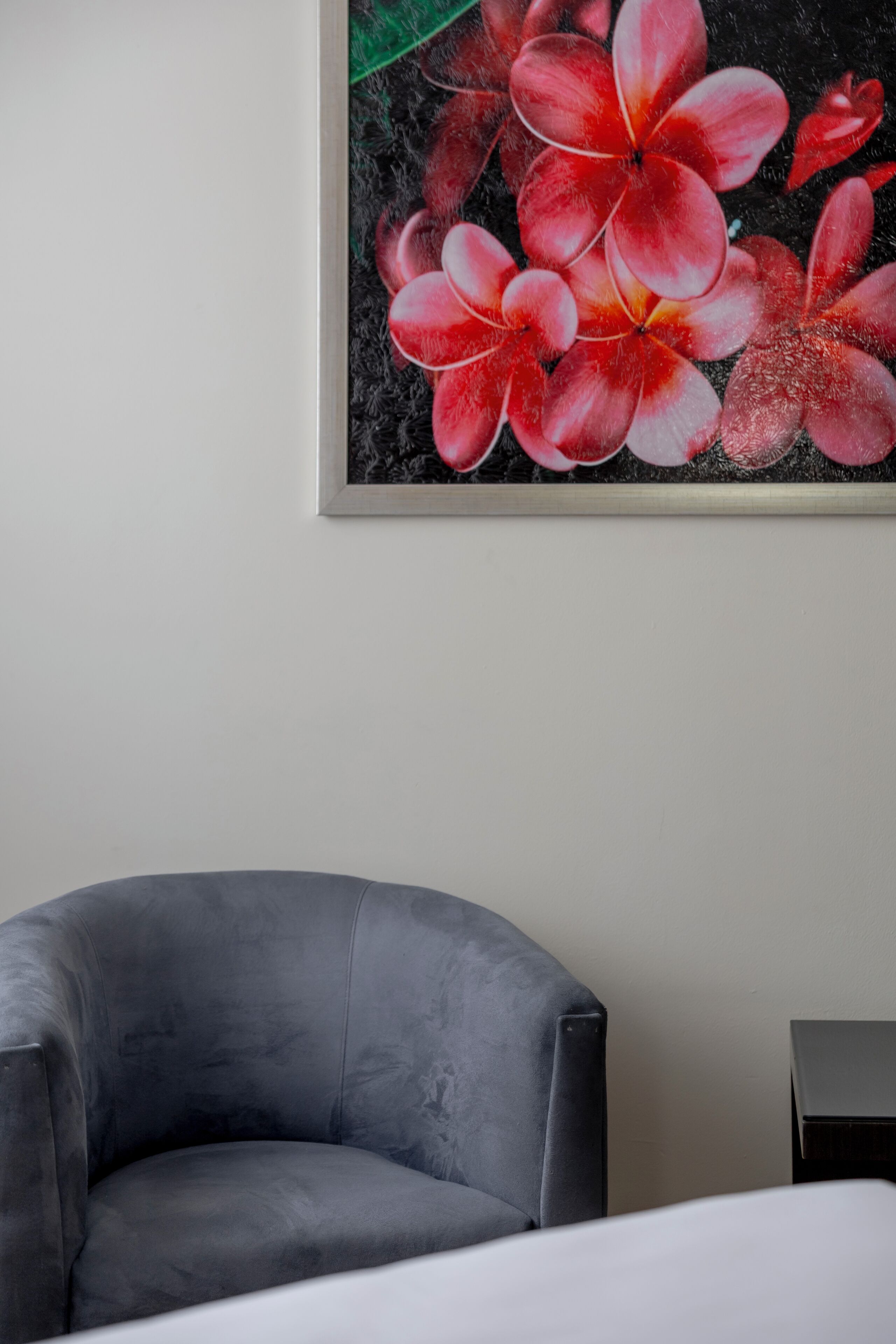 Ein Zimmer mit einem grauen Sessel, einem Bett mit weißer Bettwäsche und einem Gemälde von roten Blumen an der Wand.