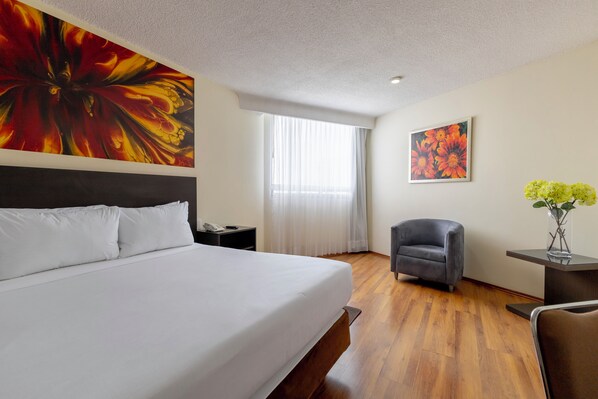 A hotel room with a large bed, a desk with a vase of flowers, a chair, and two paintings on the walls.