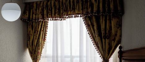 Quarto de hotel com uma cama, mesa de cabeceira, luminária pendente e janela com cortinas.