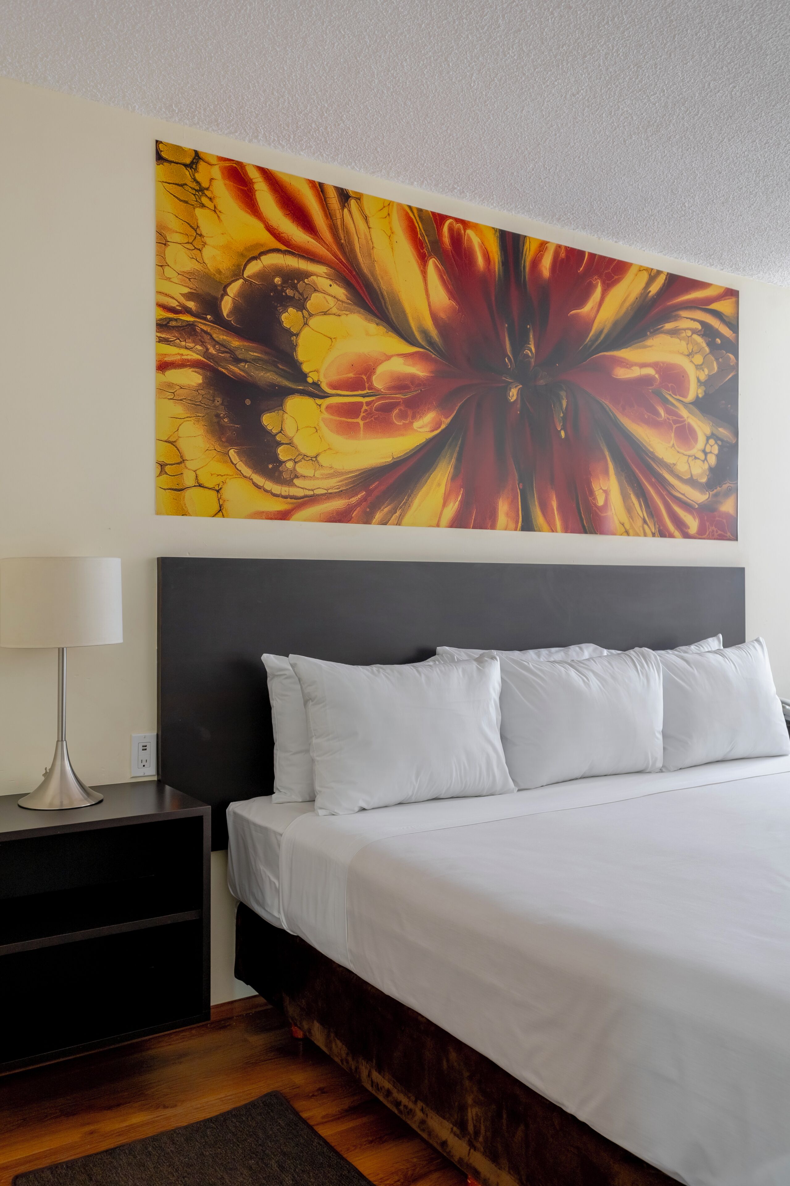 Habitación de hotel con una cama grande, una mesita de noche, una lámpara y un cuadro abstracto llamativo en la pared.