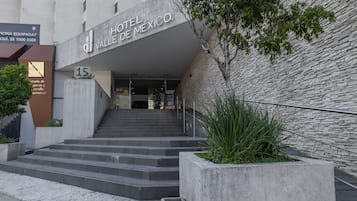 Entrada de um hotel com design moderno, apresentando uma escadaria que leva à entrada, uma grande placa acima da porta e uma área ajardinada com plantas.