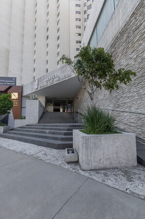 The entrance to a hotel with a modern design, featuring a staircase leading up to the entrance, a large sign above the door, and a landscaped area with plants. - Hotel Valle de Mexico Toreo (Naucalpan)