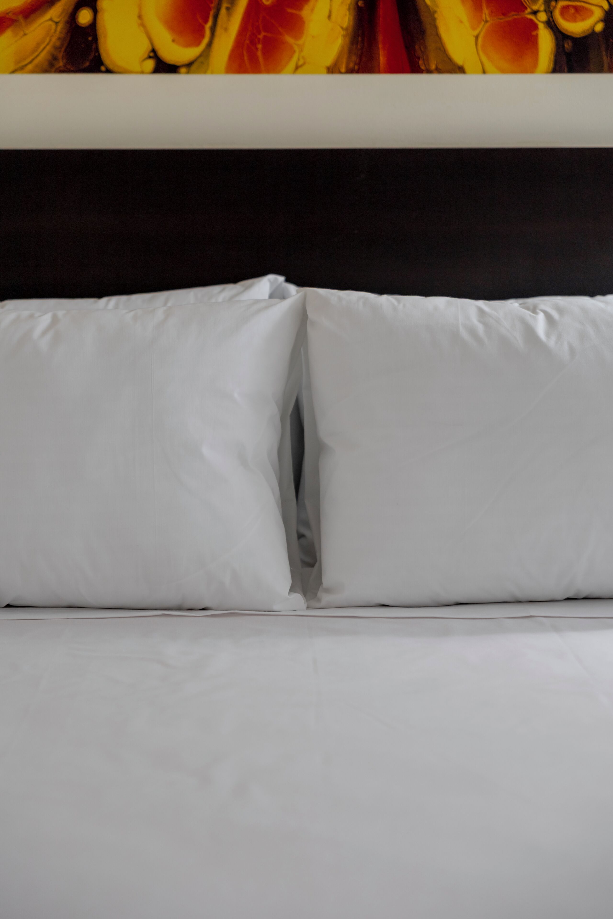 Una cama con ropa de cama y almohadas blancas, cabecera oscura y un cuadro sobre la cama.