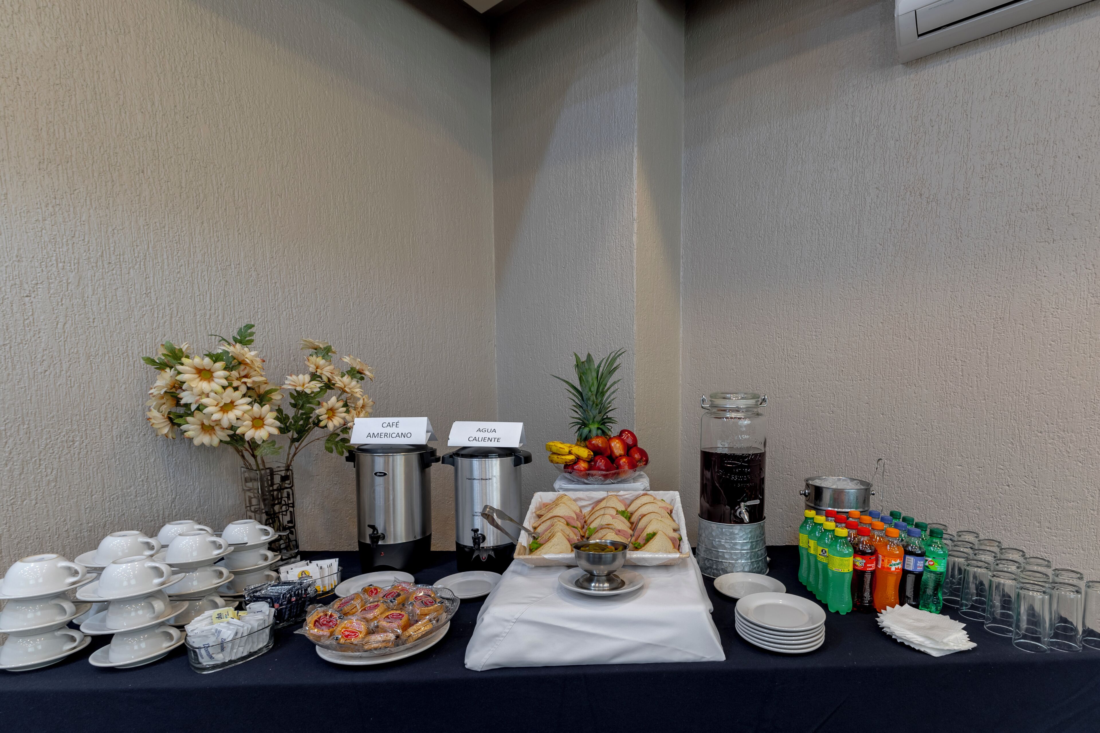 Montaje tipo bufé con dispensadores de café y té, un frutero, sándwiches y diversas bebidas.