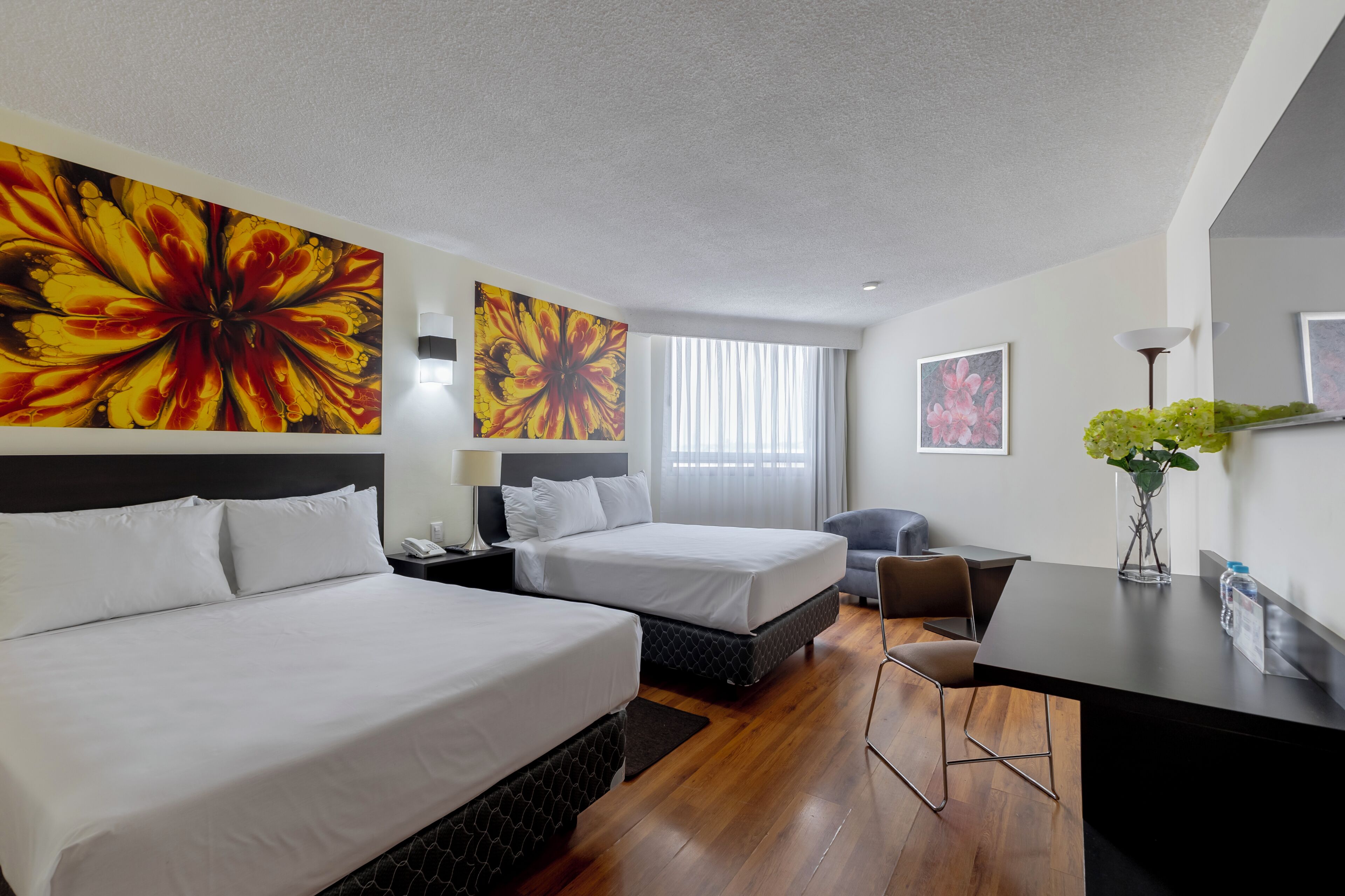 Een hotelkamer met twee bedden, een bureau, een stoel en grote bloemrijke kunst aan de wanden.