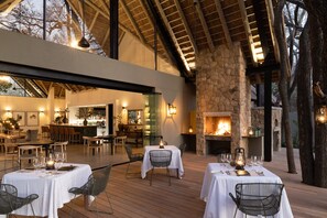 An outdoor dining area with tables set for two, a stone fireplace, and a bar area visible through a glass partition. - Monwana (Hoedspruit)