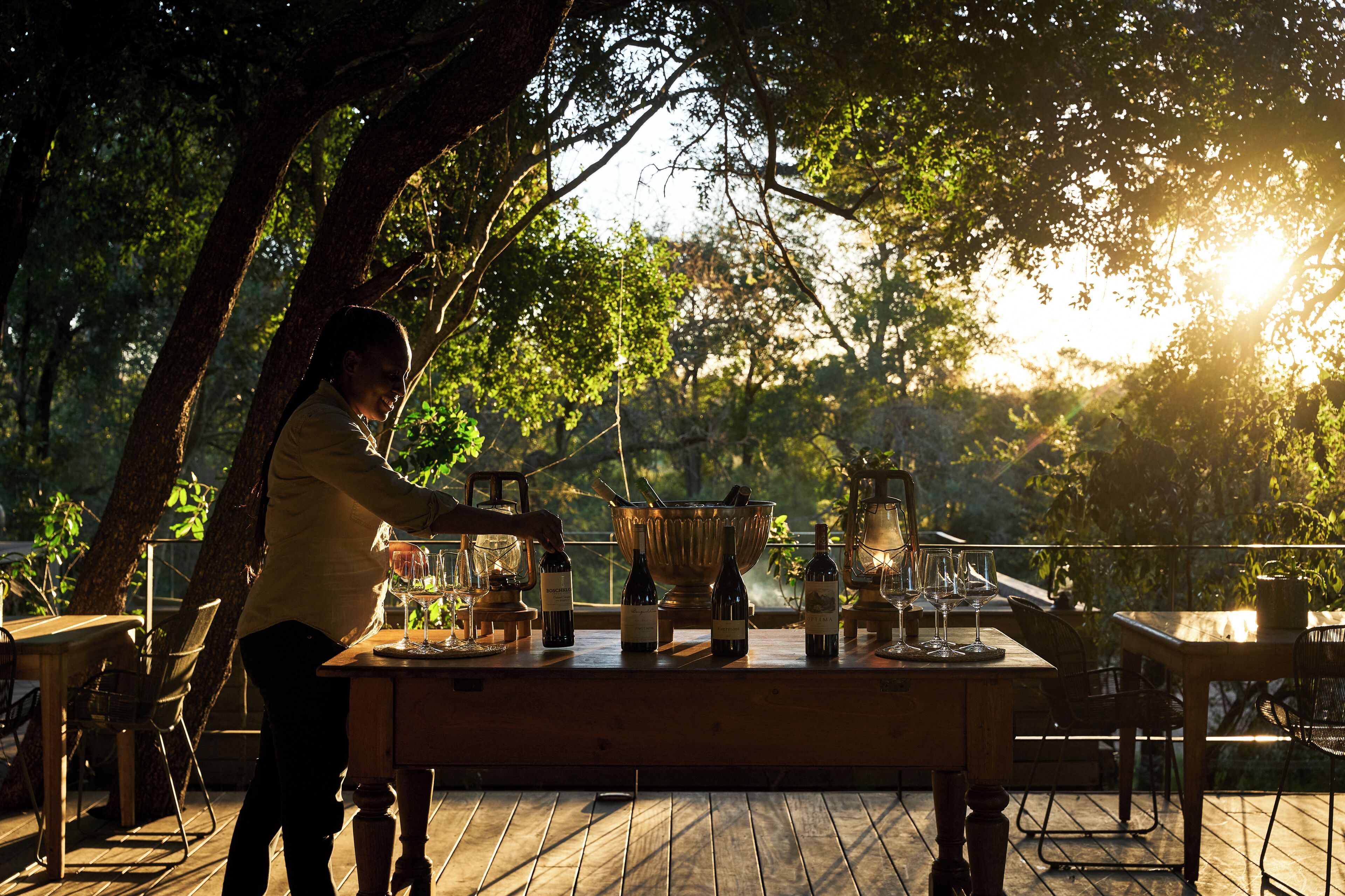 An outdoor dining area with a wooden table set for a meal, surrounded by trees and natural light.