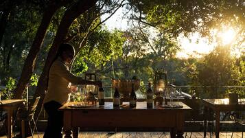 An outdoor dining area with a wooden table set for a meal, surrounded by trees and natural light.