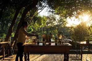 Un espace de restauration en plein air avec une table en bois dressée pour un repas, entourée d’arbres et baignée de lumière naturelle.