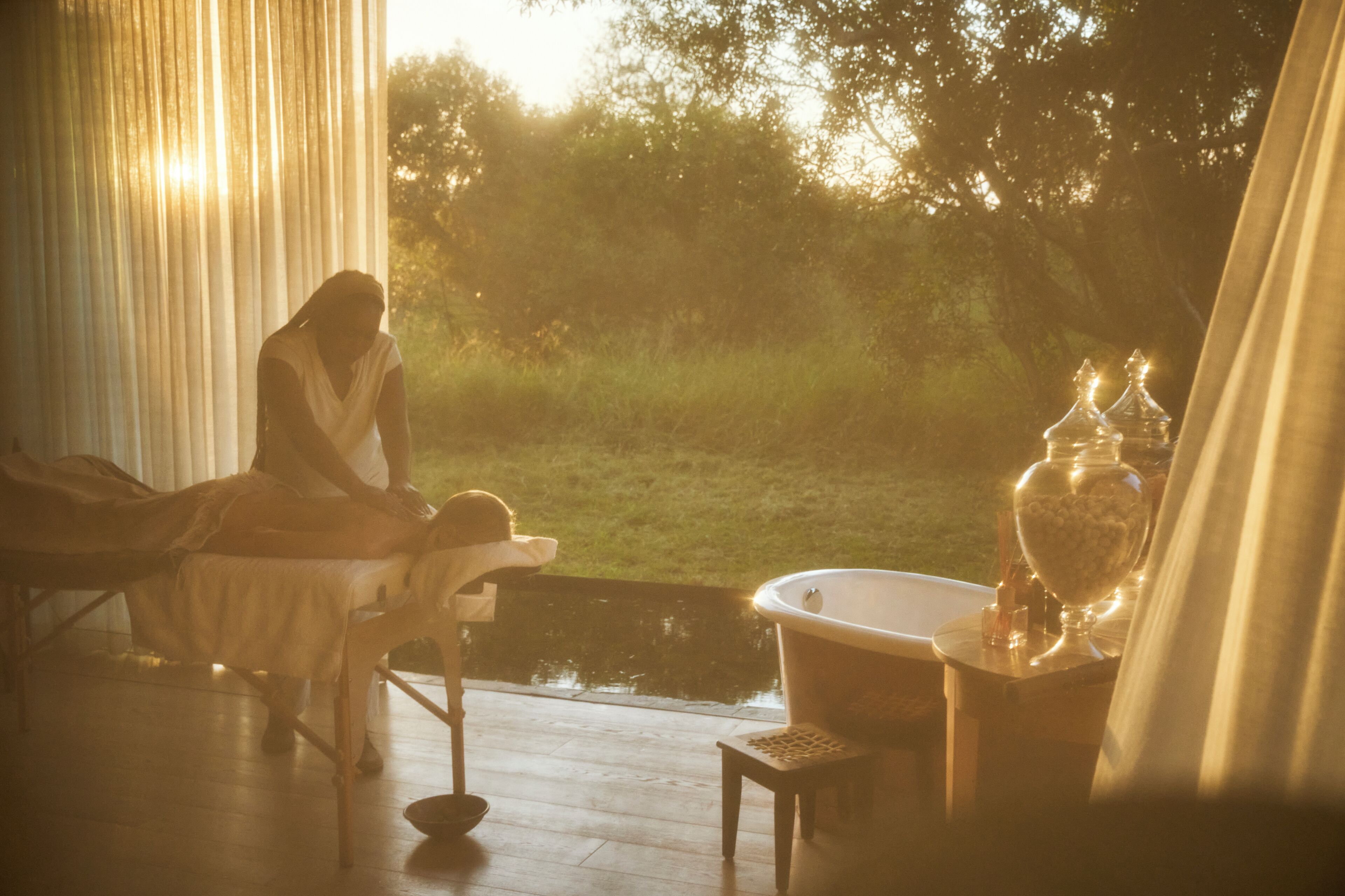 A room with a bed, a small table with a lamp, and a view of a garden.