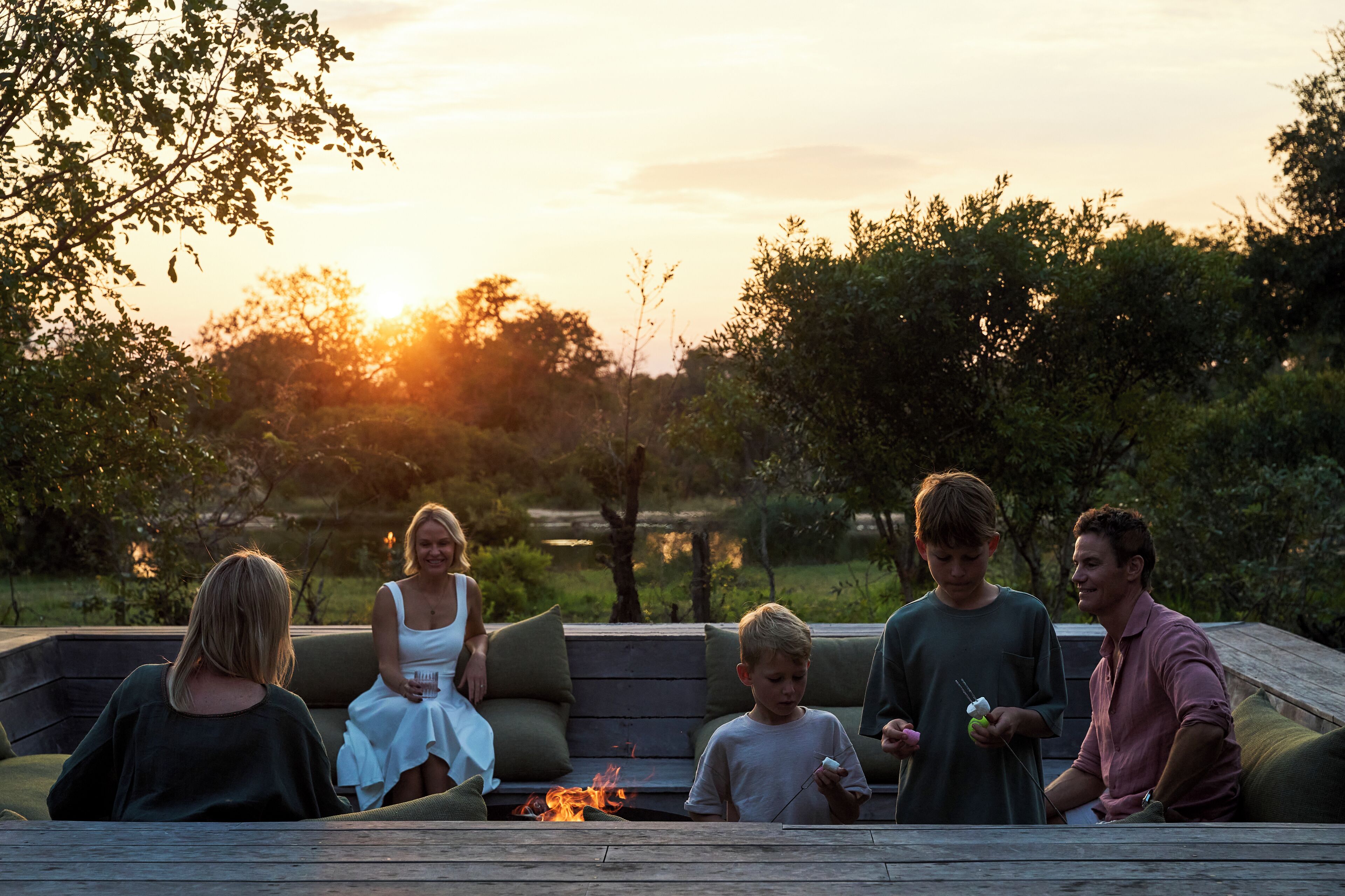 A group of people sitting on a wooden deck with a fire pit in the center, surrounded by greenery and trees.
