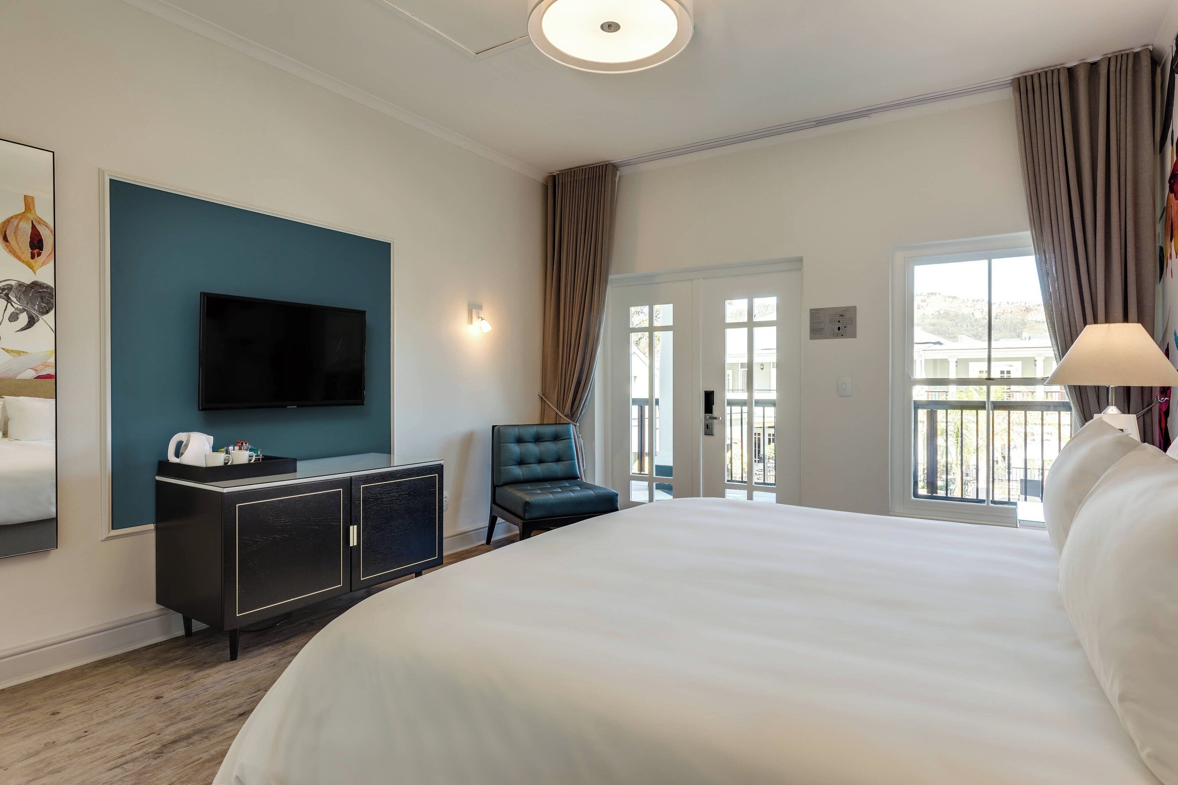 Une chambre d’hôtel avec un grand lit, un téléviseur à écran plat fixé au mur, un meuble de couleur sombre contenant une machine à café, un fauteuil bleu et une vue sur l’extérieur par une porte.