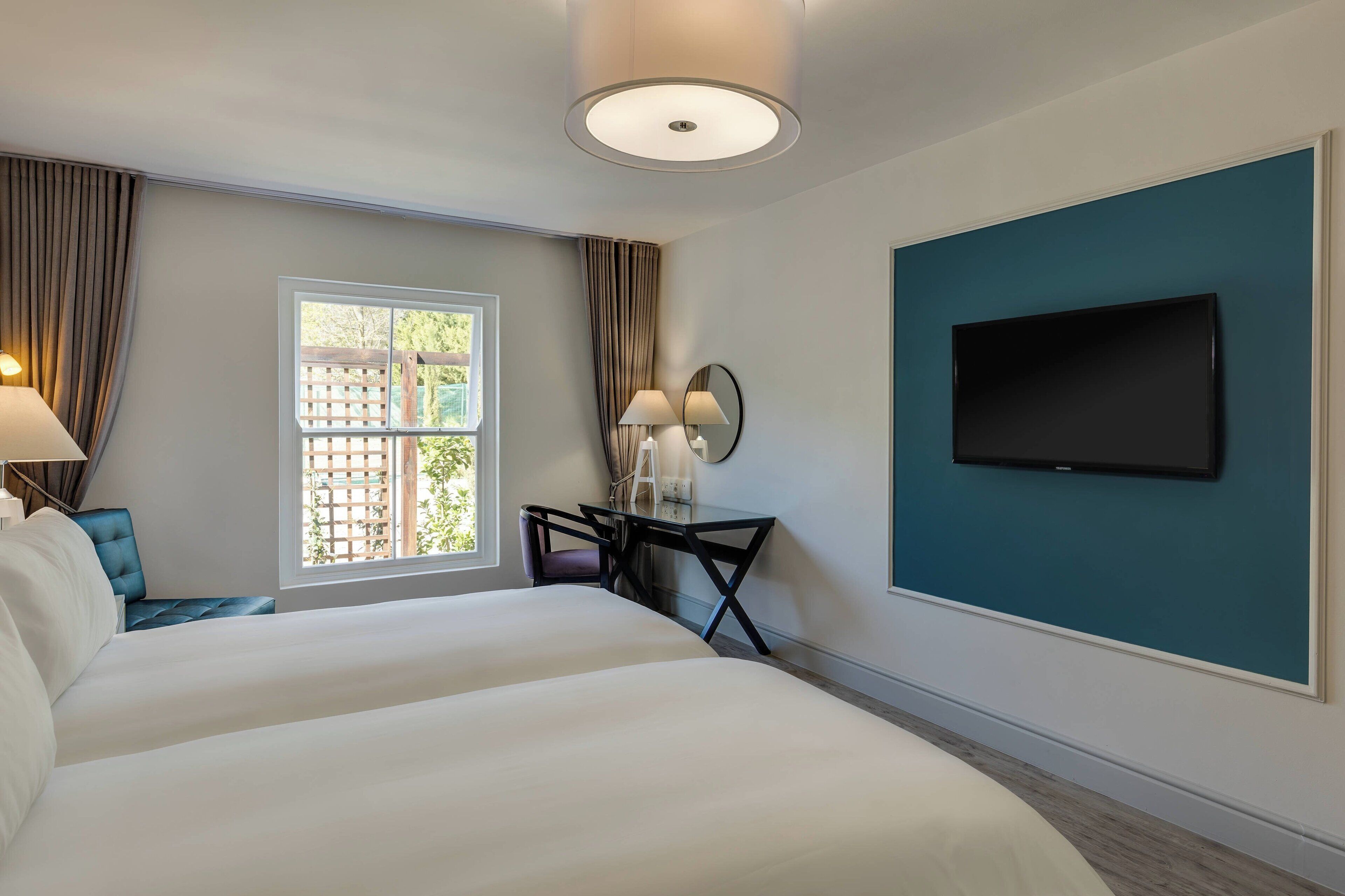 A hotel room with a large bed, a desk with two lamps, a TV mounted on the wall, and a window with curtains.