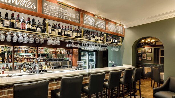 A bar area with a long counter, multiple shelves of bottles, and several black bar stools.