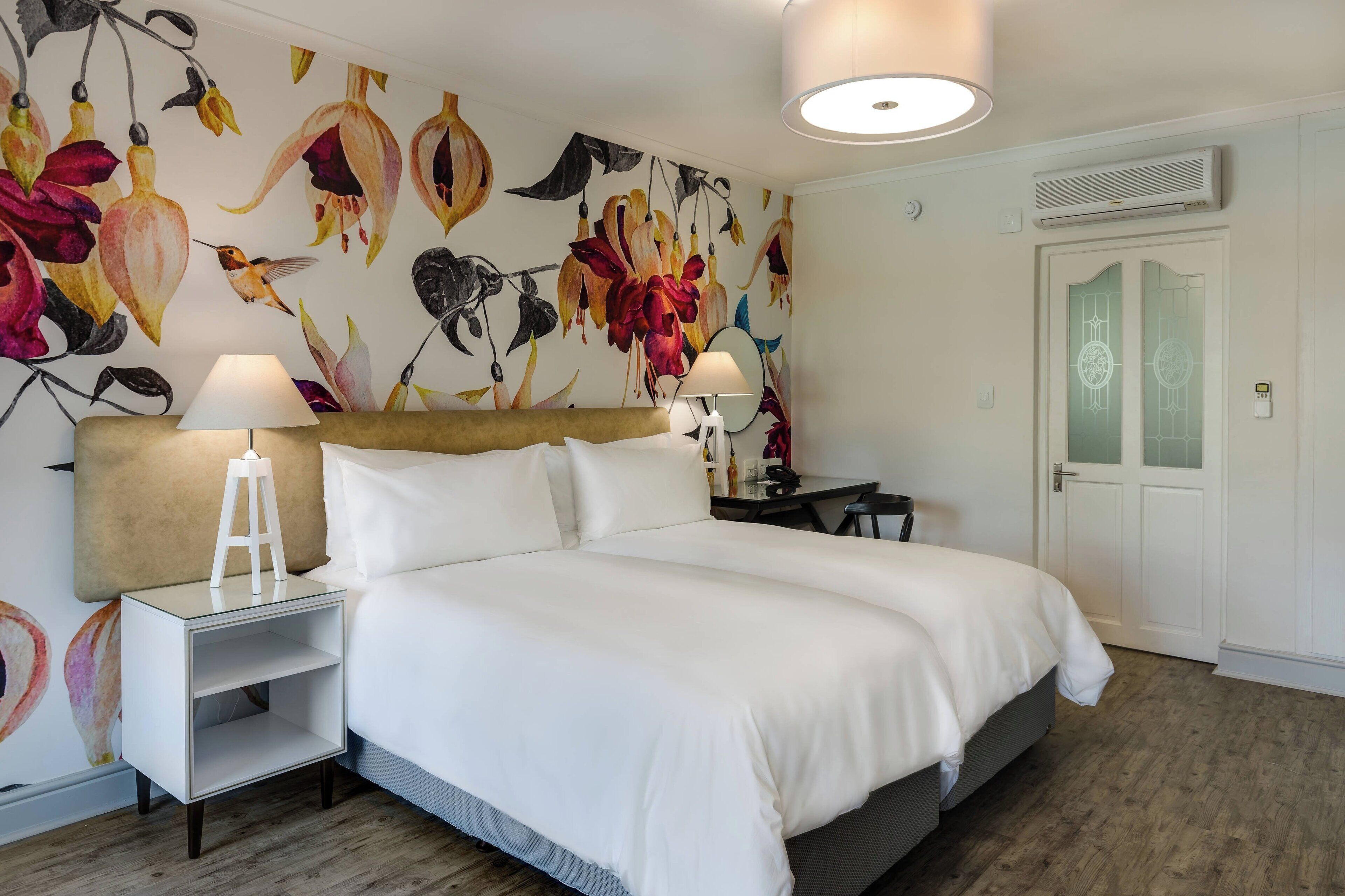 A bedroom with a bed, two bedside tables, a nightstand, a wall with a floral mural, a door with frosted glass panels, and a ceiling light.