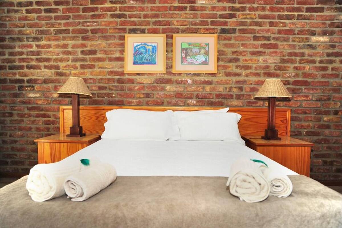 A hotel room with a brick wall, a wooden bed, two nightstands with lamps, and two framed pictures on the wall.