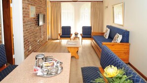 Una habitación acogedora con pared de ladrillo, un sofá de madera, dos sillones, una mesa de centro y un televisor montado en la pared.