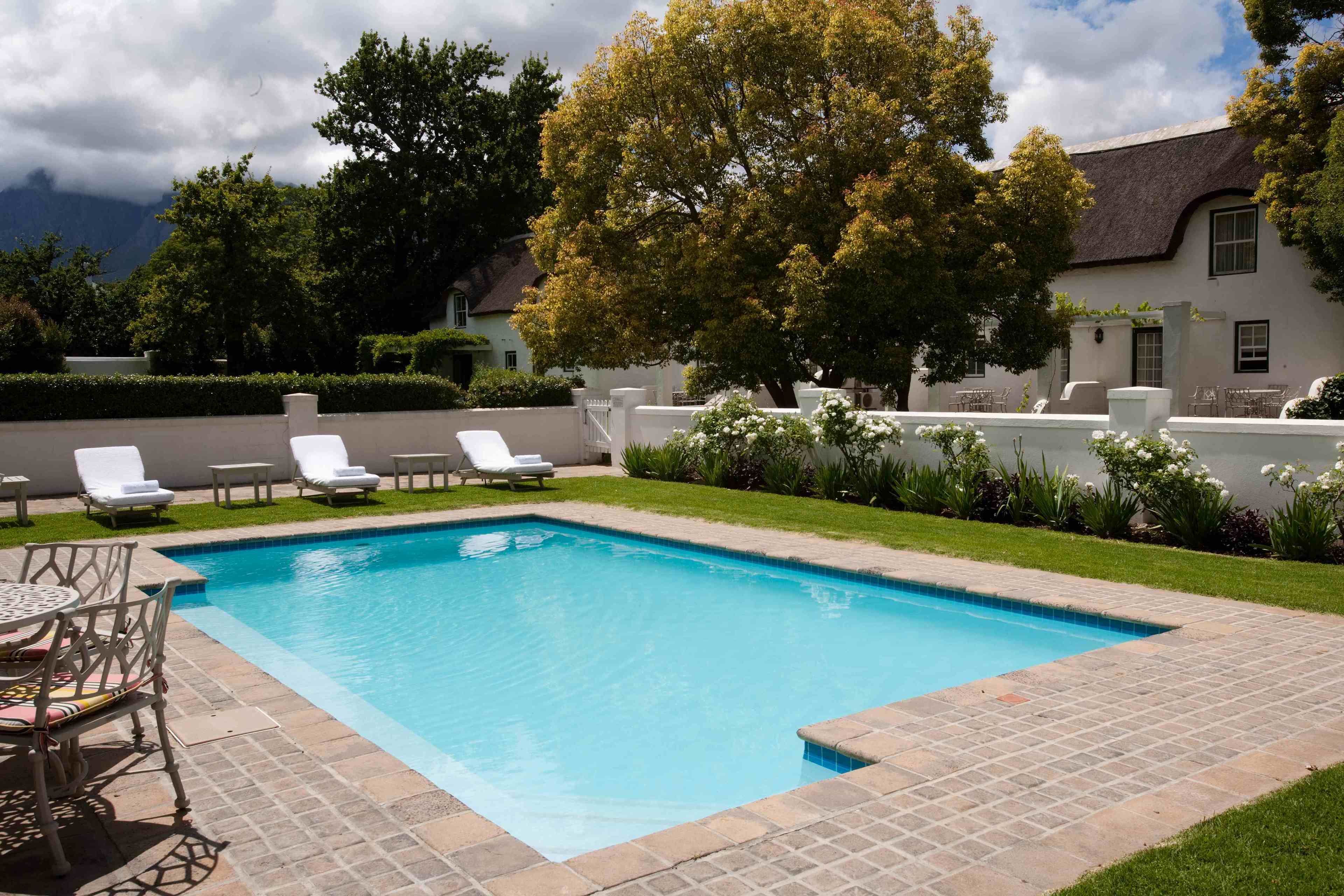 A rectangular swimming pool with a tiled edge, surrounded by a grassy area and a white wall. There are white lounge chairs and a table with chairs along the pool's edge.
