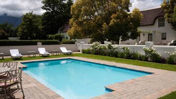 A rectangular swimming pool with a tiled edge, surrounded by a grassy area and a white wall. There are white lounge chairs and a table with chairs along the pool's edge.