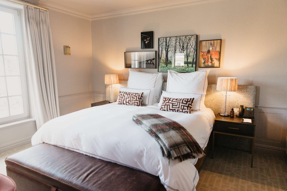 A neatly made bed with white linens and patterned pillows, a bedside table with a lamp, and a window with curtains.