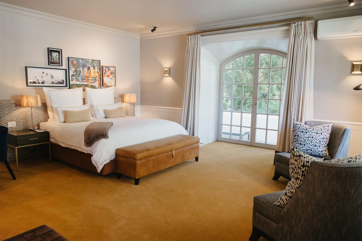 A spacious bedroom with a large bed, a sitting area with a chair, and a view of greenery through a large window.
