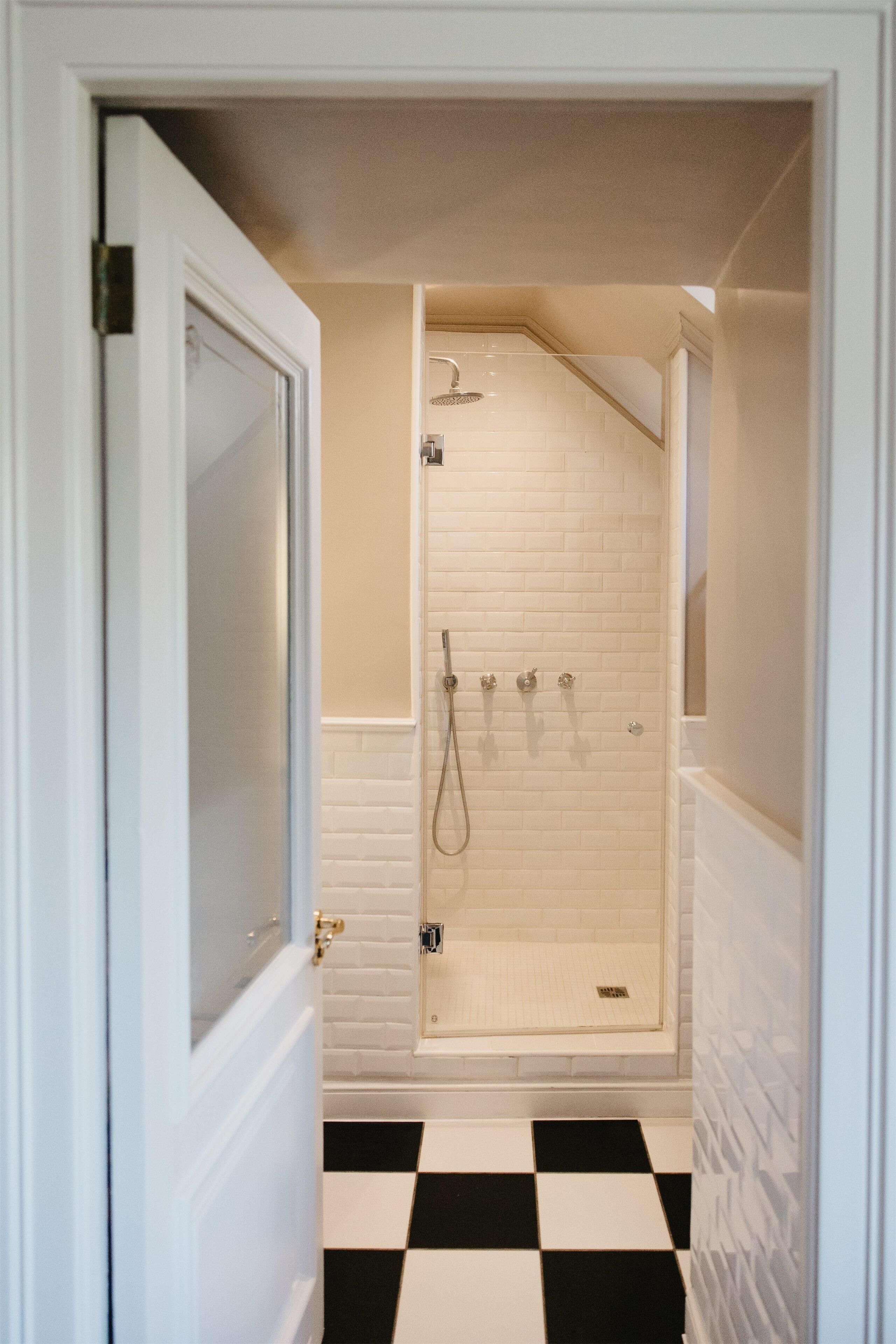 Un couloir étroit mène à une salle de bain dont le sol est à carreaux et qui est équipée d’une douche avec un mur recouvert de carreaux blancs.