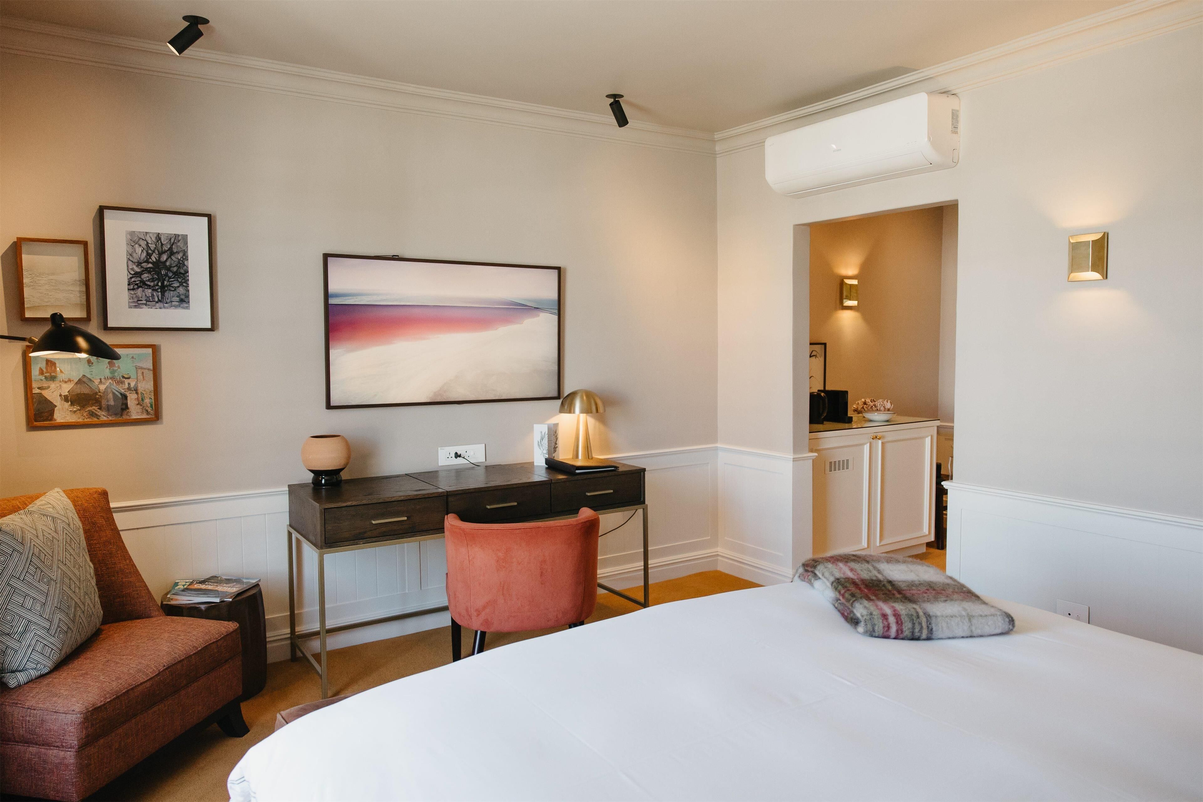 A hotel room with a bed, a desk, a chair, a small table, and a nightstand. There is a painting on the wall, a lamp on the desk, and a window with a view of the outside.