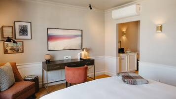 A hotel room with a bed, a desk, a chair, a small table, and a nightstand. There is a painting on the wall, a lamp on the desk, and a window with a view of the outside.
