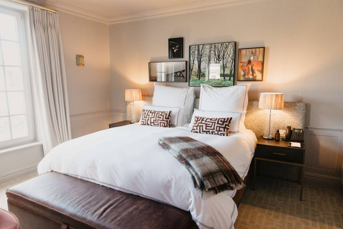 A neatly made bed with white linens and patterned pillows, a bedside table with a lamp, and a window with curtains.