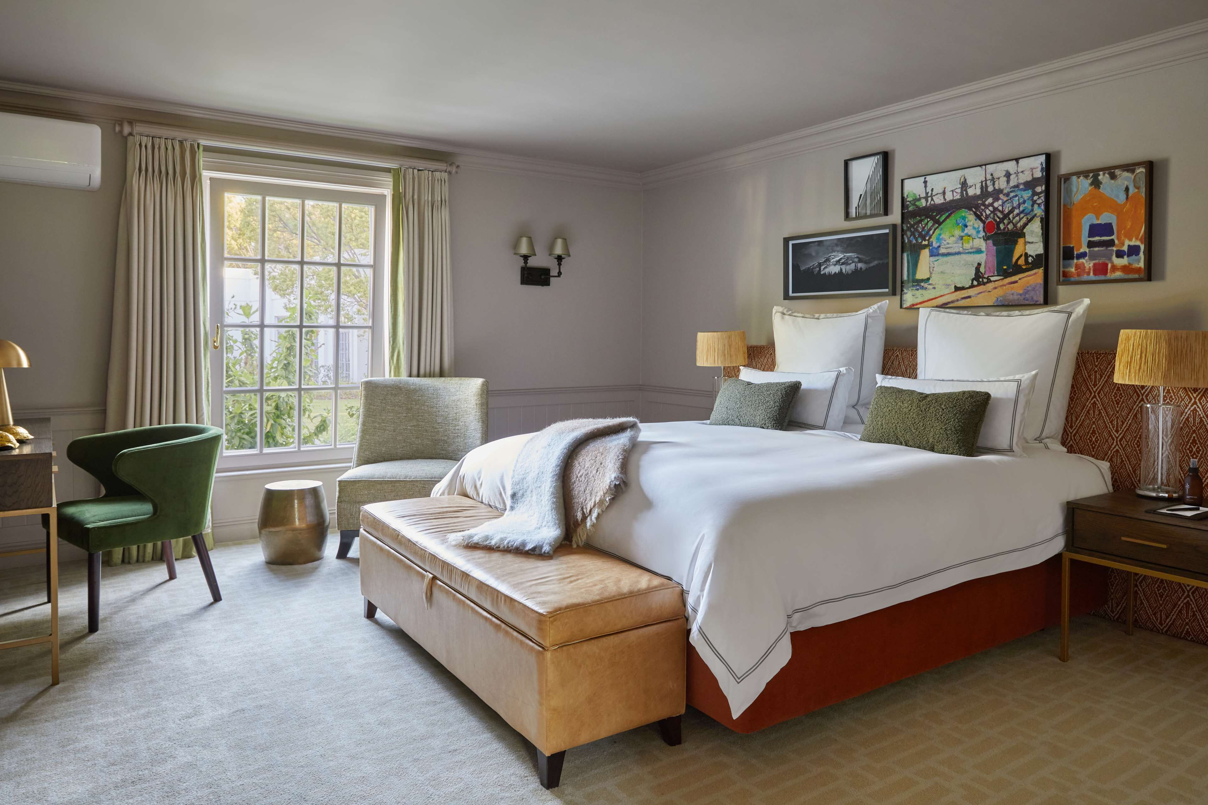 A hotel room with a large bed, two chairs, a bench, a desk, and a window with curtains.