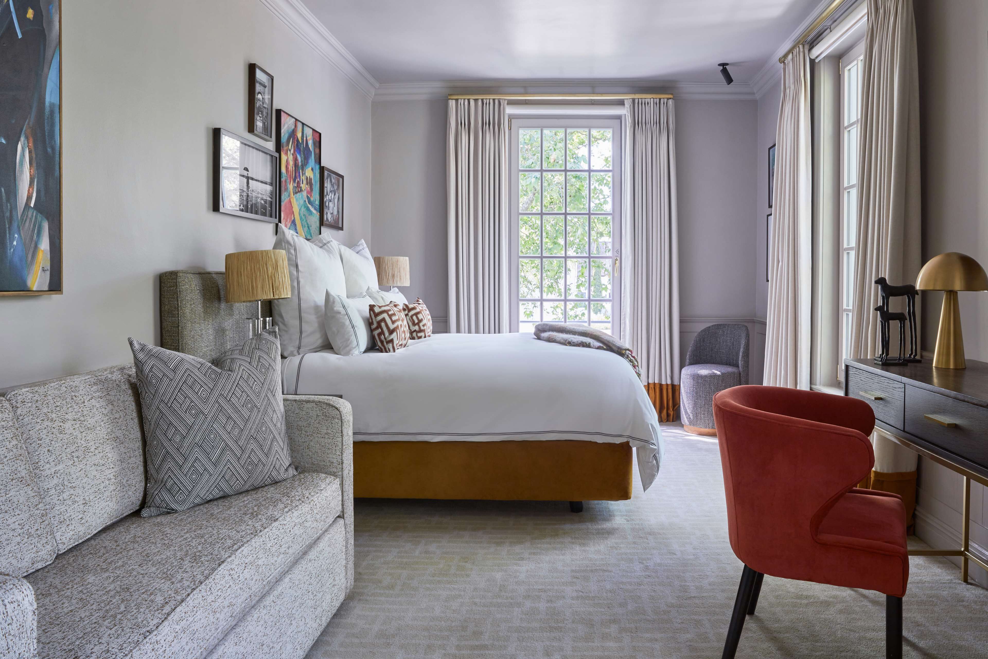 A bedroom with a bed, a grey sofa, a red chair, and a desk with a lamp.