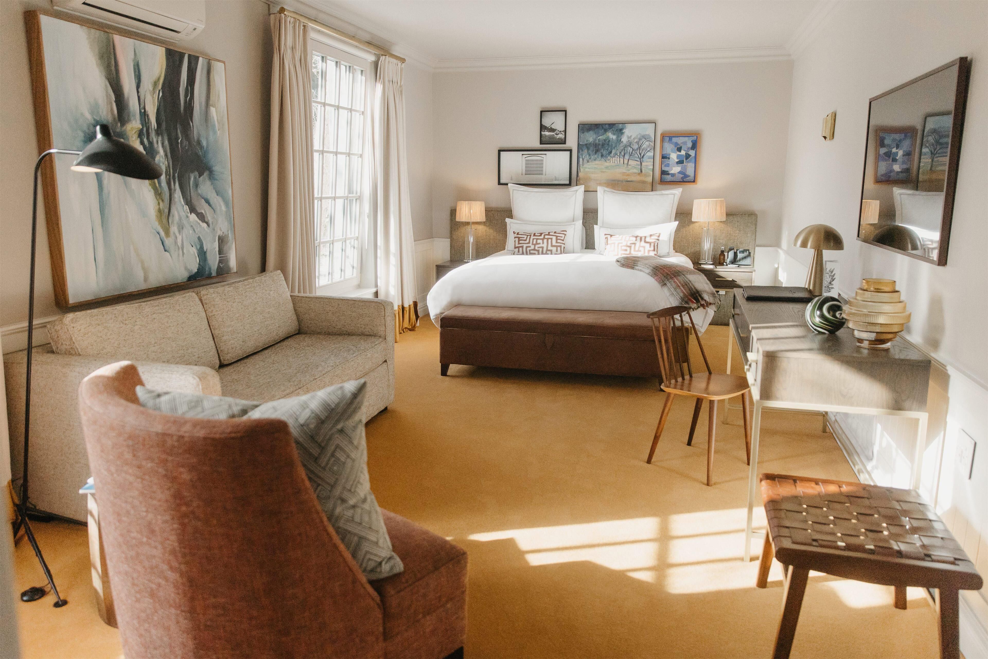 A hotel room with a large bed, a sofa, a desk, and a chair. There are two lamps on the desk and a large painting on the wall.